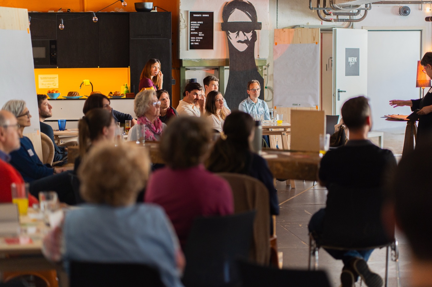 Gemeinschaftsabend in Unsere Tischlerei in Melk — Teilnehmer:innen sitzen an Tischen in der offenen Werkstatt, im Hintergrund Küche und ein Mural, eine Sprecherin steht.